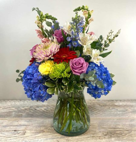 A blooming assortment of blue hydranga, spider mums, lavender roses, alstroemeria, blue delphinium and pink snap dragon arranged in a glass conch vase.